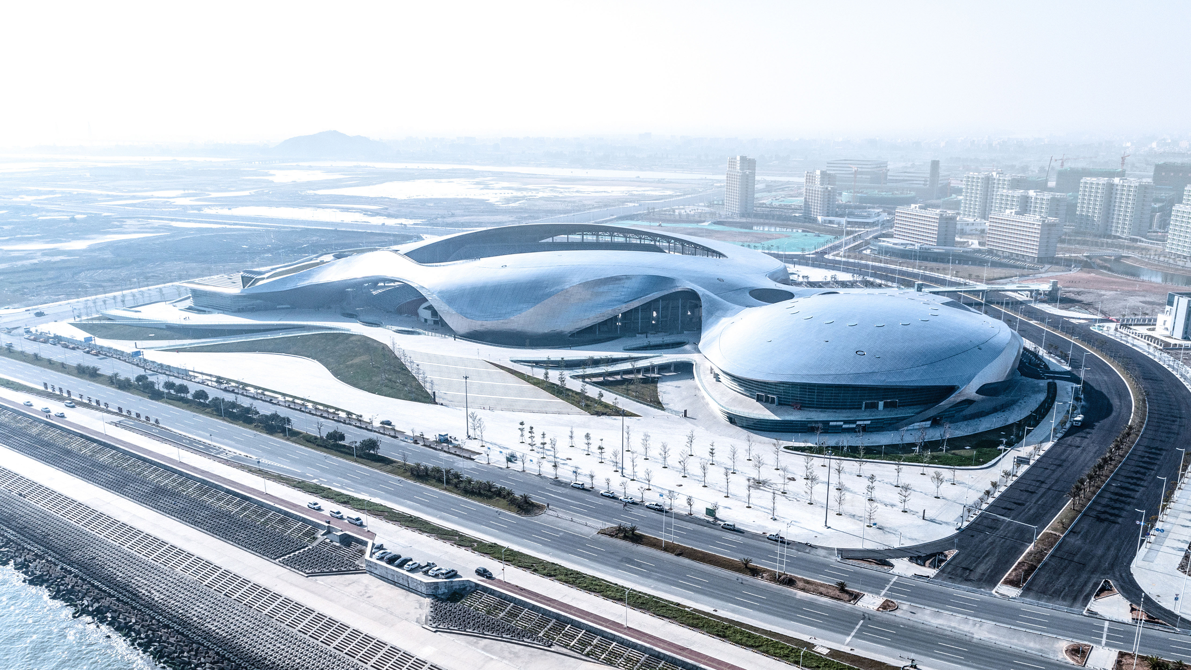 Daytime aerial view of the East Campus of Shantou University and Asian Youth Games Venue, showing the stadium and gymnasium roof forms against the coastline.